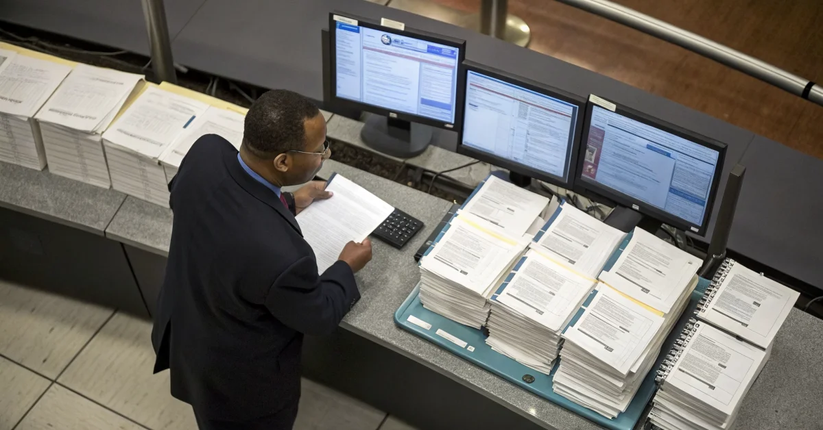 Government official reviewing household documents and data records at desk, computer screens visible, administrative office s