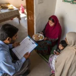 Pakistani household survey enumerator with clipboard interviewing family in modest home, formal documentation process, wide a